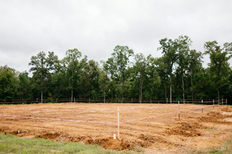 Freshly graded building lot with string lines and stakes, tree line behind